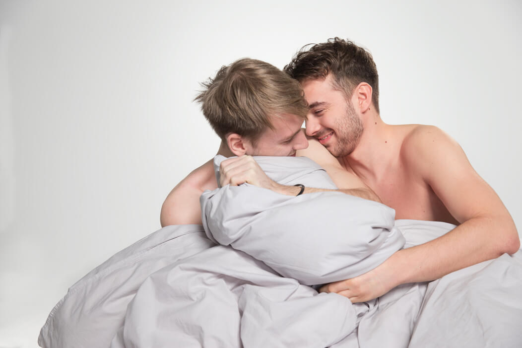 Schwules Paar kuschelt und albert unbekleidet unter der Bettdecke, beide lachen sich an.